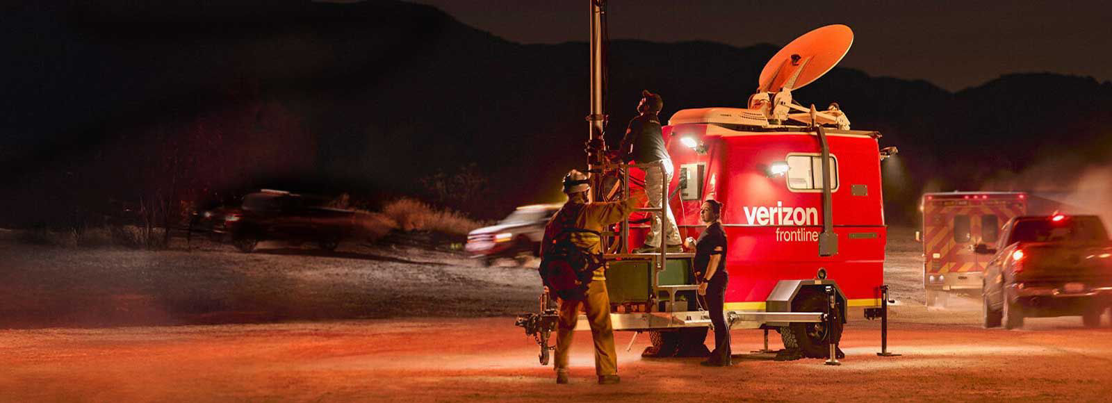 Emergency responders working around a lit-up red Verizon Frontline trailer with a satellite dish during a nighttime operation.